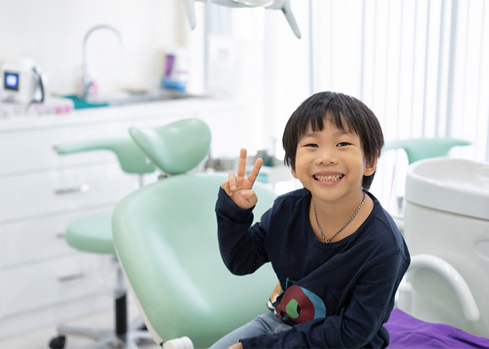 image of a little boy sitting on a dental chair holding up a peace sign and smiling